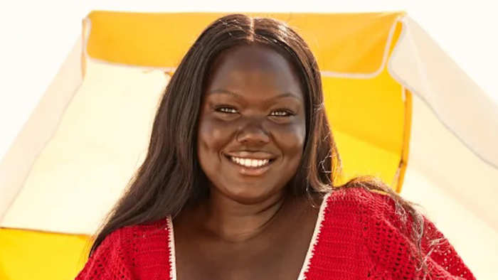 Nyma Tang stands in front of a yellow and white beach umbrella in a red knit swimsuit and smiles at the camera.
